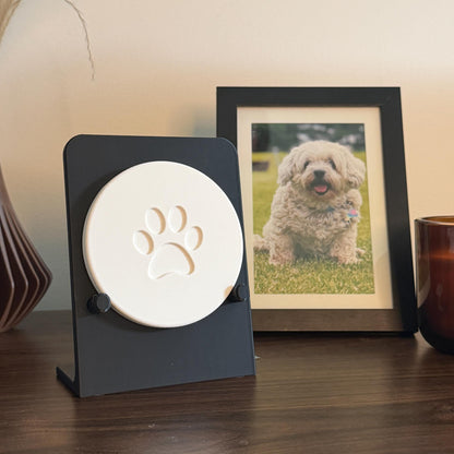 Black Essential paw print holder displaying a large white round sample with a stylized paw design, beside a framed dog photo.