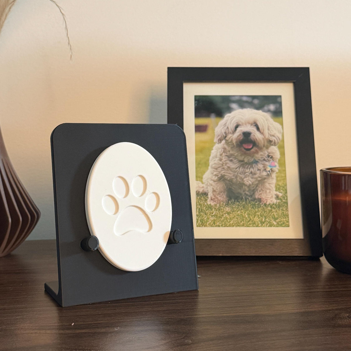 Black Essential paw print holder displaying a small white oval sample with a stylized paw design, beside a framed dog photo.