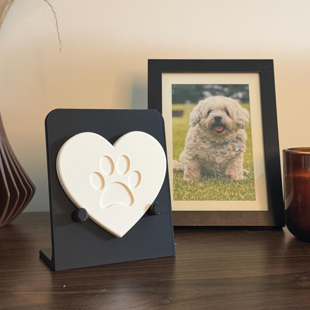 Black Essential paw print holder displaying a white heart-shaped sample with a stylized paw design, beside a framed dog photo.
