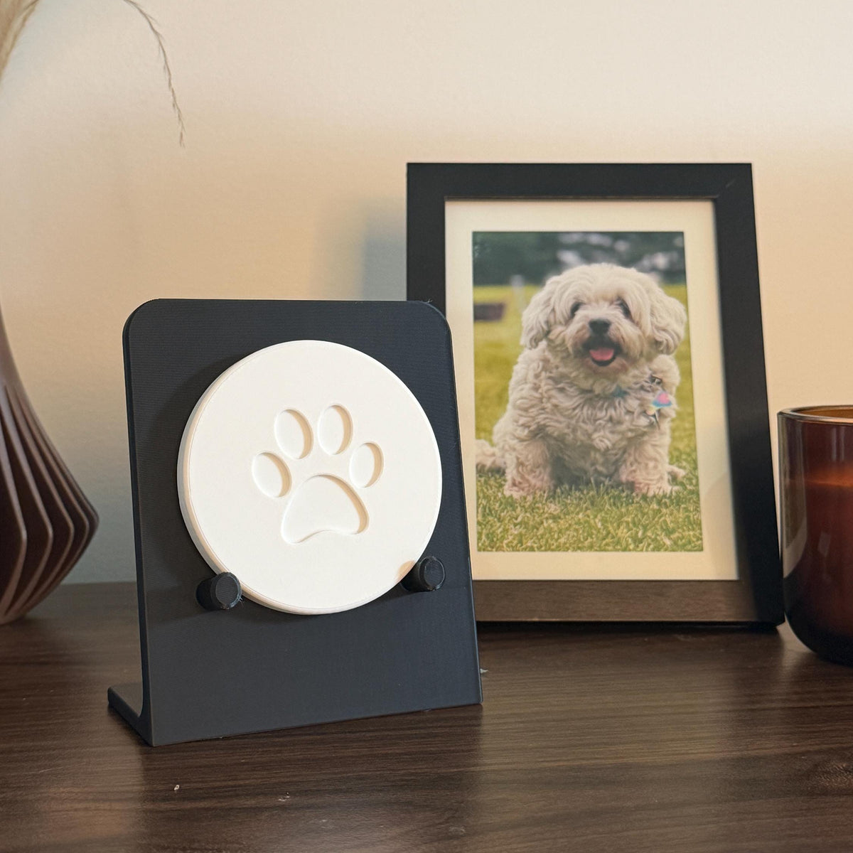 Black Essential paw print holder displaying a small white round sample with a stylized paw design, beside a framed dog photo.